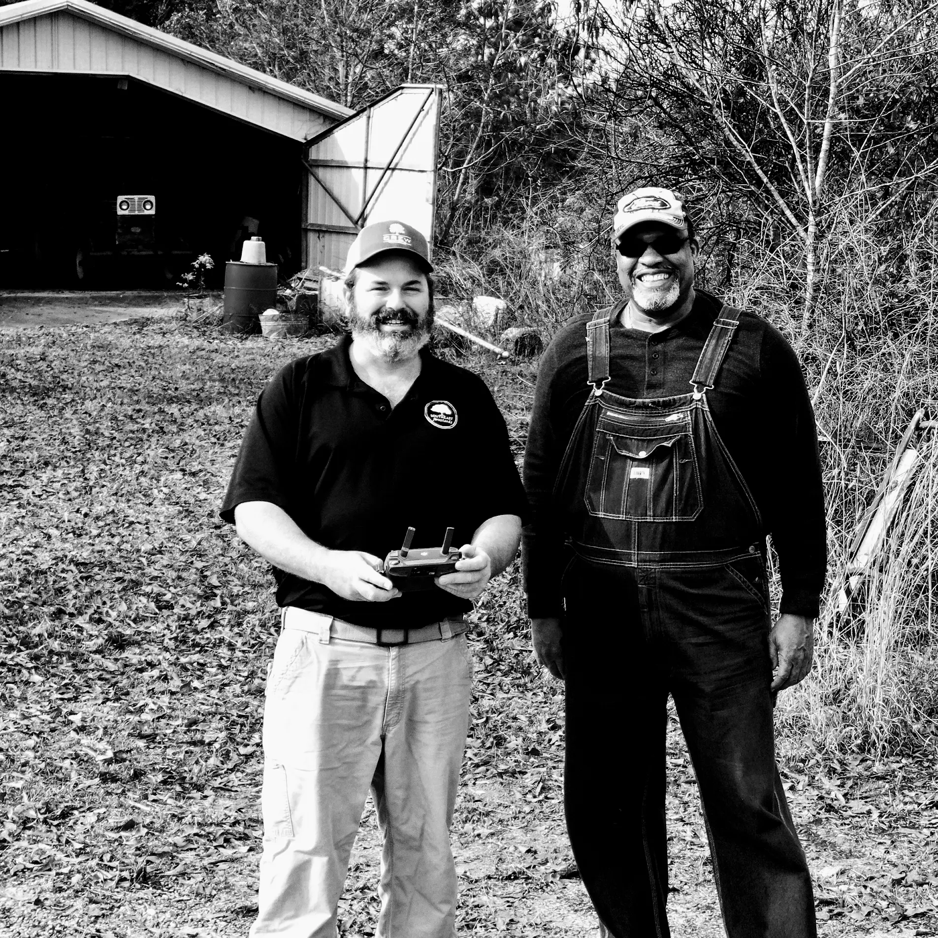 Two men standing outdoors near a wooded property, one holding a drone controller.